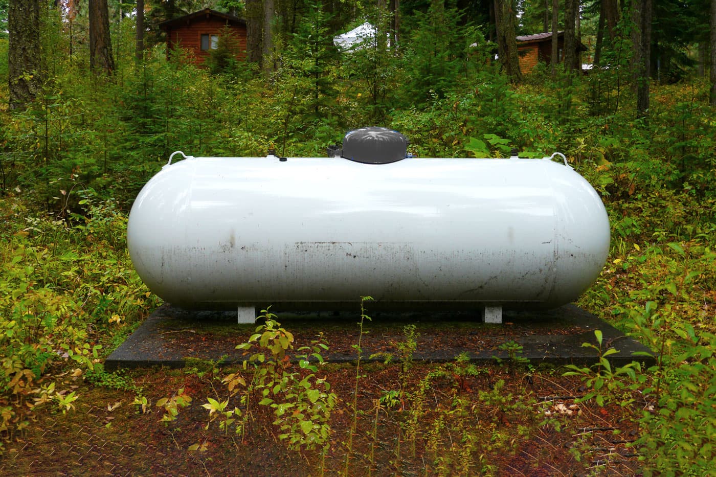 White propane tank in a forested area next to a house. 