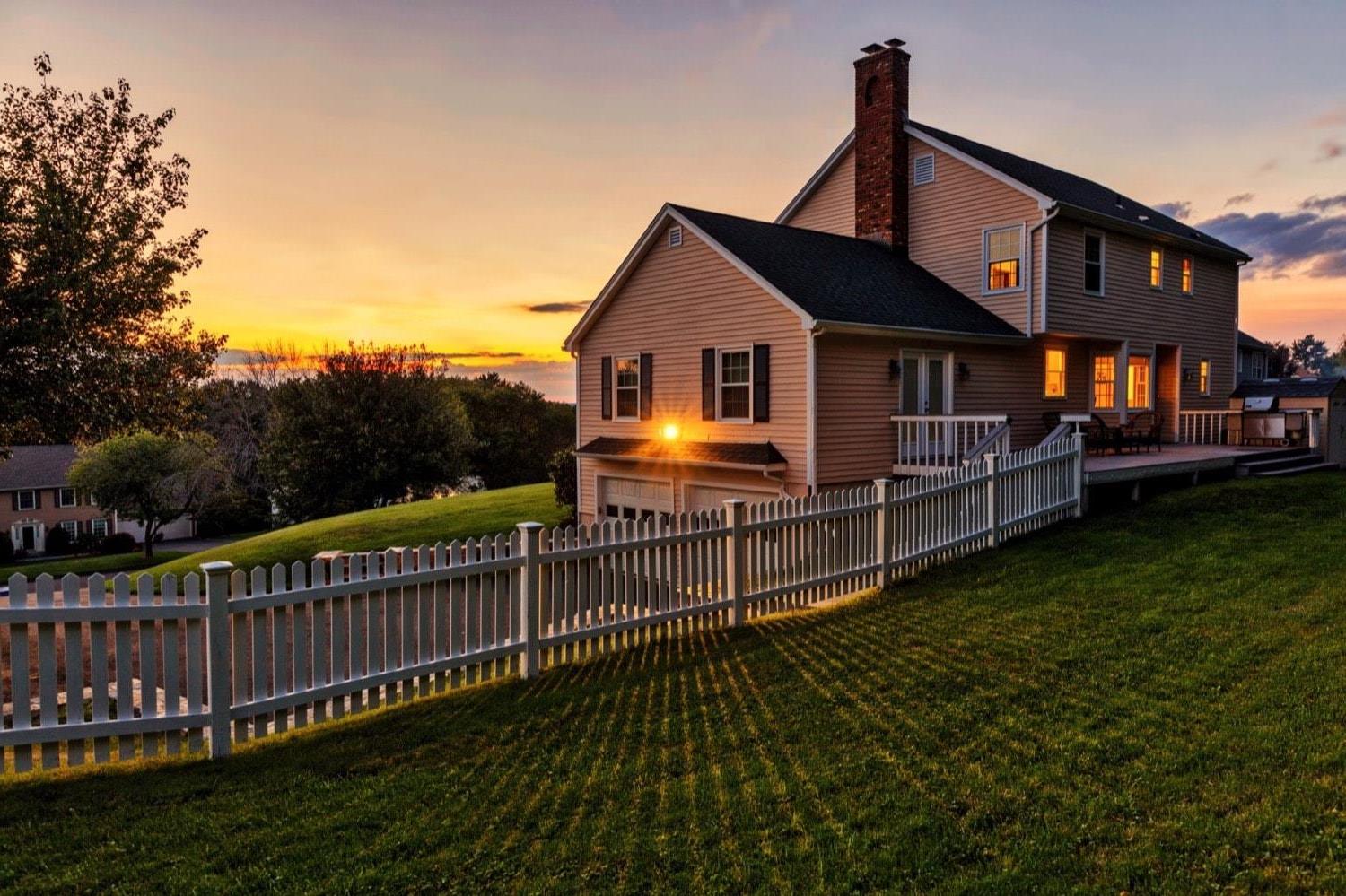 The back of a house with a back porch, BBQ, and white picket fence, at sunset.