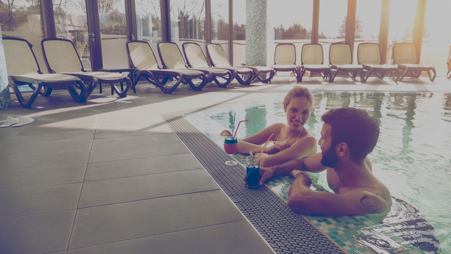 Couple at the edge of a pool, smiling and drinking cocktails. 