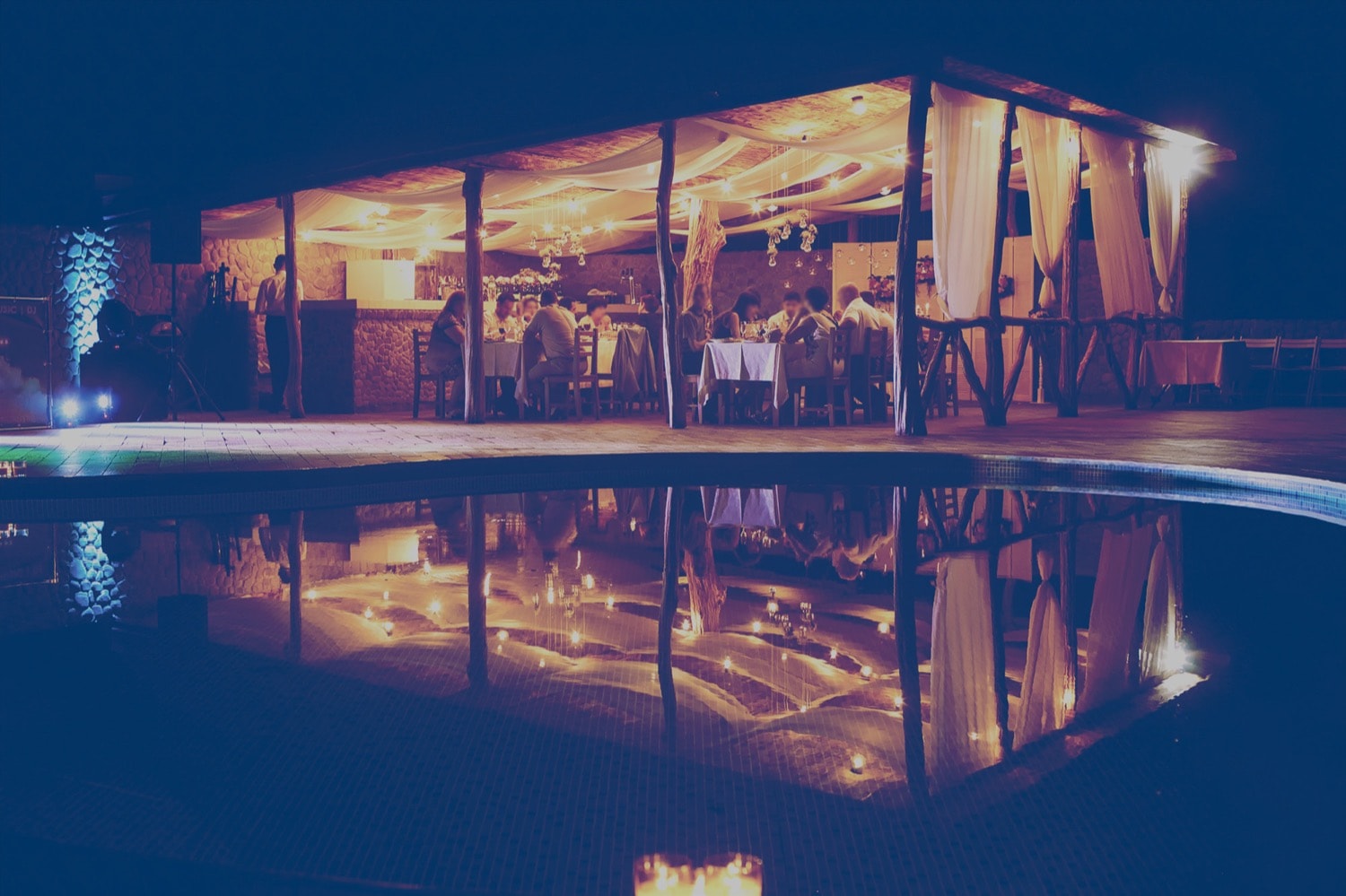 A large group of people eating a meal at night in an outdoor restaurant, next to a pool. 