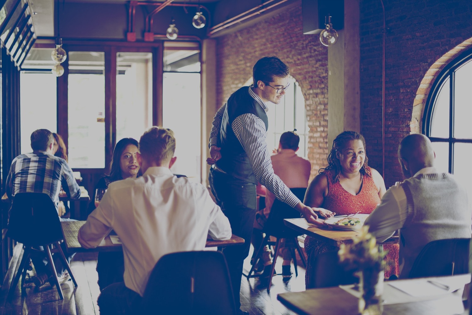 Multiple people seated in a restaurant. The server is giving a meal to a couple. 