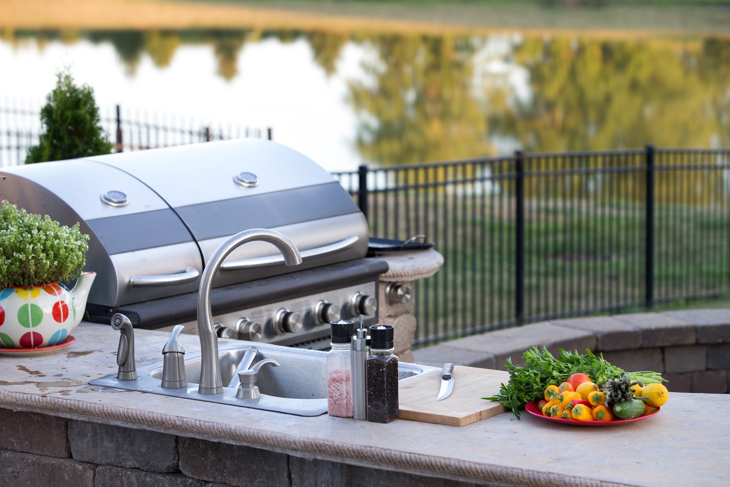 An outdoor residential kitchen with a sink and propane grill situated next to a lake. 