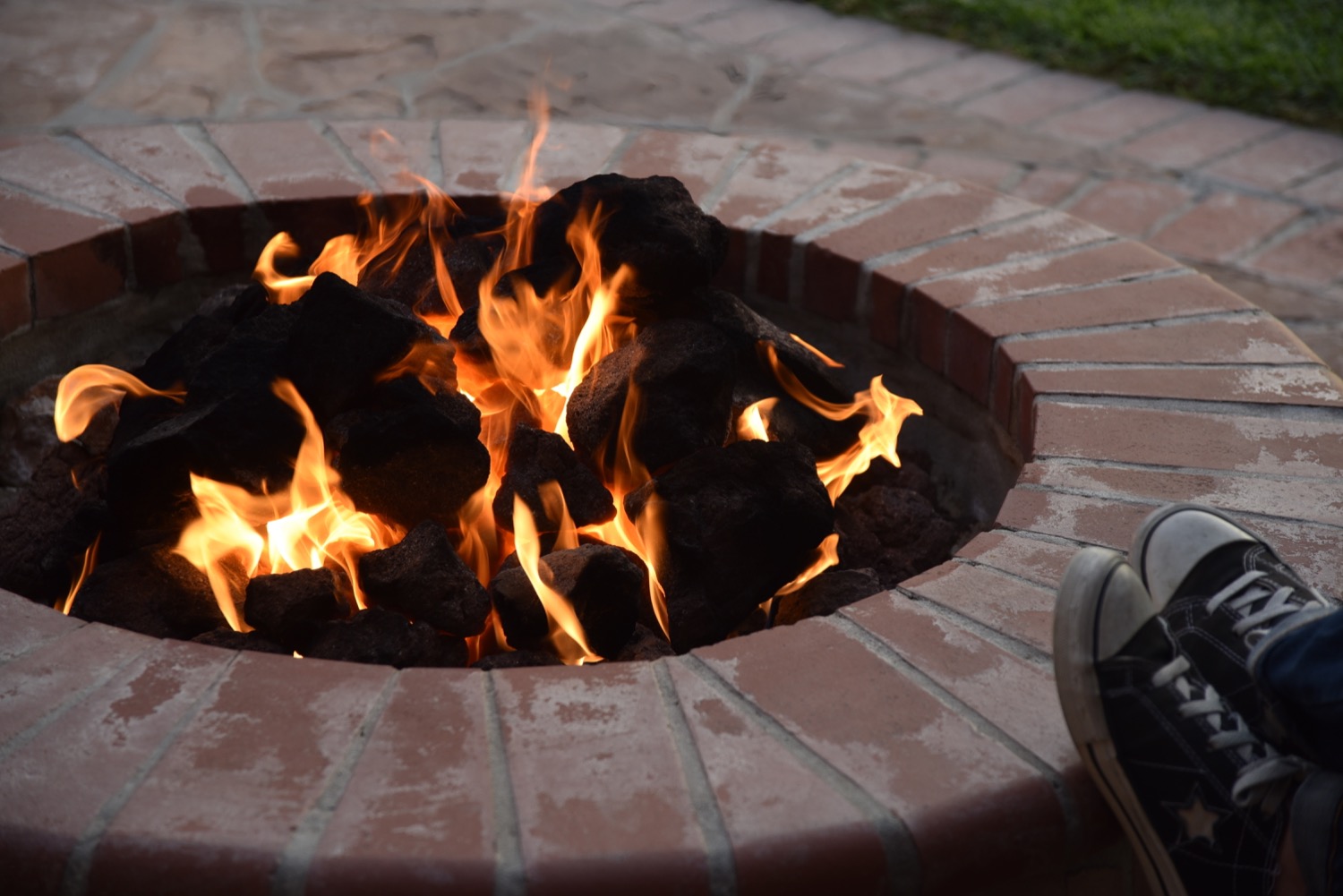 Person sitting next to a propane-powered outdoor fire pit made of brick. 